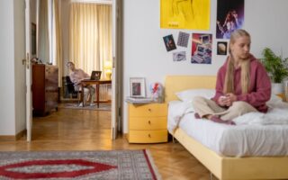 A teenage girl is sitting on a bed in her bedroom while her mom, sitting at her desk, tries to look inside from far away.