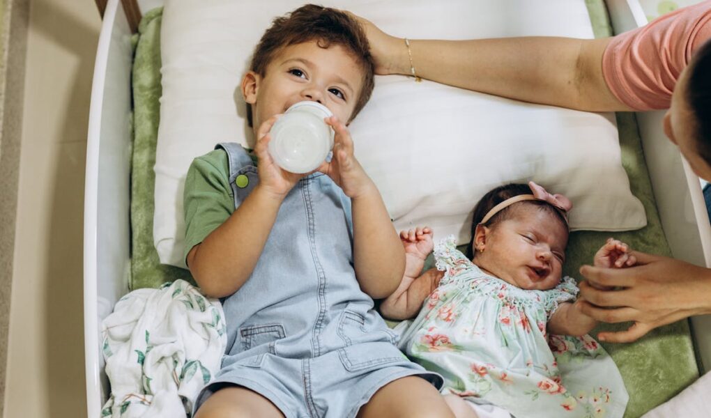 Two children laying down. Older child drinking from bottle while baby who is the second child is laying next to him while mom holds her hand and pats the older boy's head.