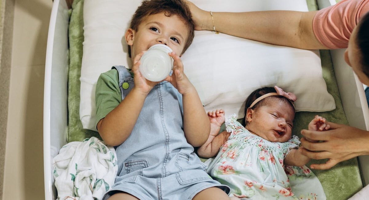 Two children laying down. Older child drinking from bottle while baby who is the second child is laying next to him while mom holds her hand and pats the older boy's head.