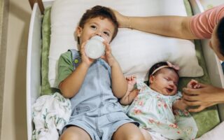 Two children laying down. Older child drinking from bottle while baby who is the second child is laying next to him while mom holds her hand and pats the older boy's head.