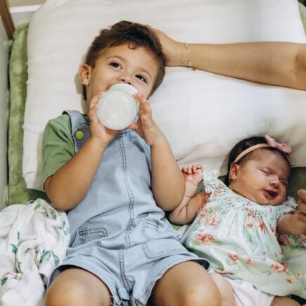 Two children laying down. Older child drinking from bottle while baby who is the second child is laying next to him while mom holds her hand and pats the older boy's head.