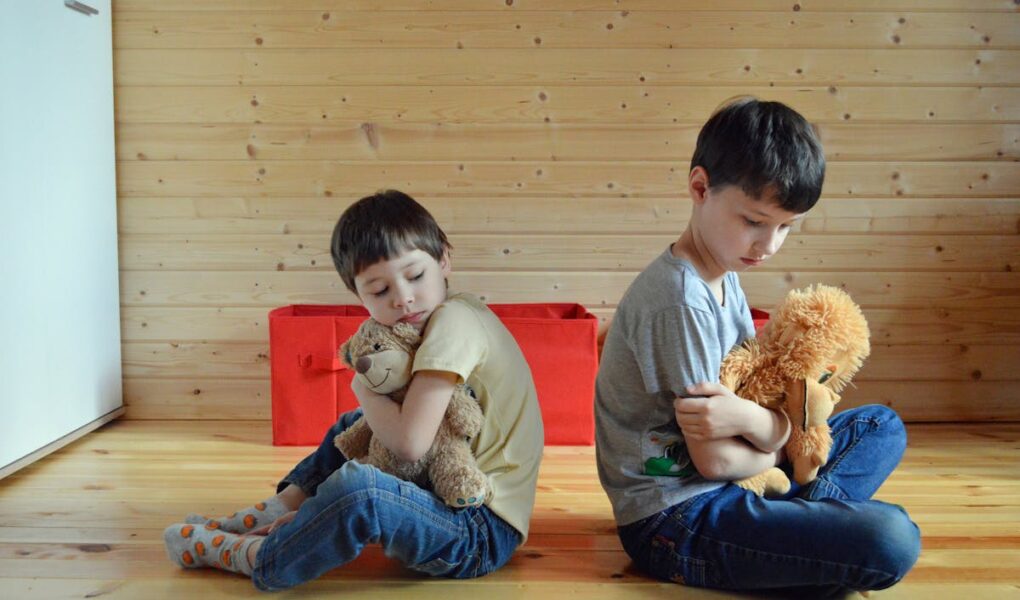 Two children hugging teddy bears are sitting down backs turned on each other. The two boys seem to be upset with each other.