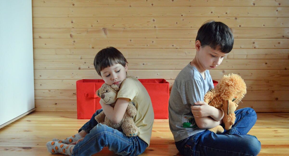 Two children hugging teddy bears are sitting down backs turned on each other. The two boys seem to be upset with each other.