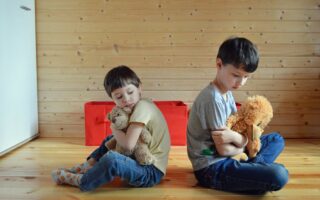 Two children hugging teddy bears are sitting down backs turned on each other. The two boys seem to be upset with each other.