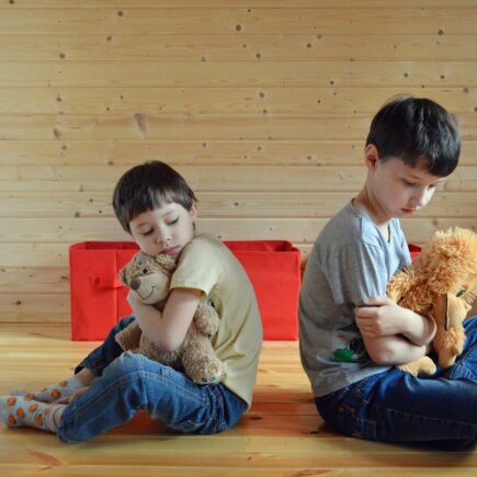 Two children hugging teddy bears are sitting down backs turned on each other. The two boys seem to be upset with each other.