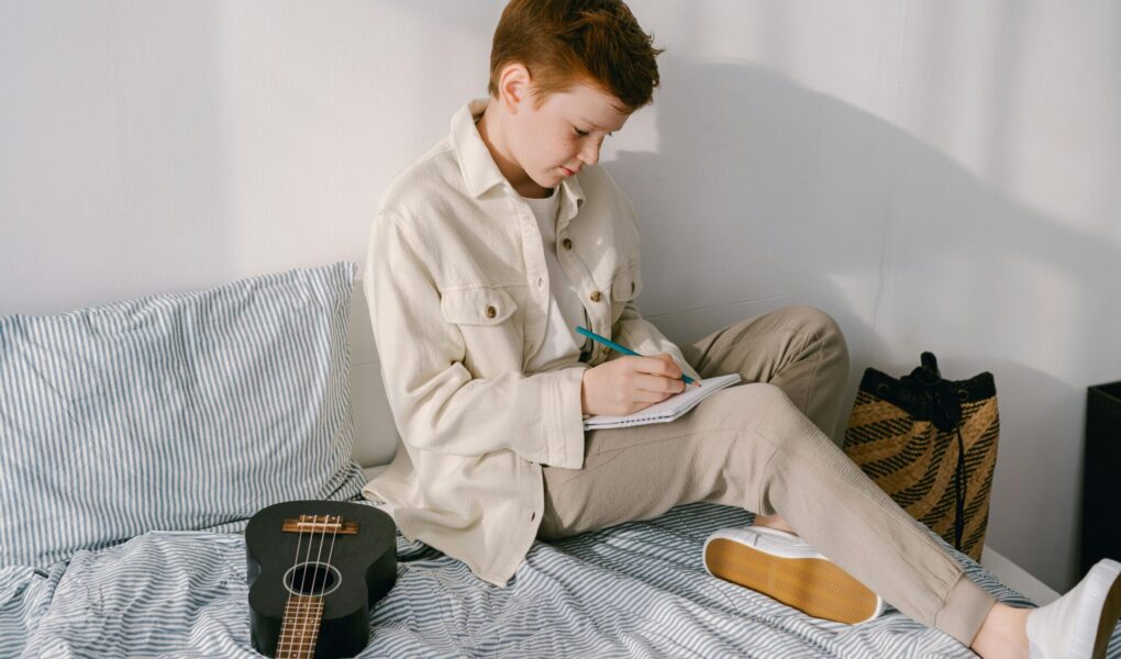 Preteen boy sitting on his bed writing on a piece pf paper.