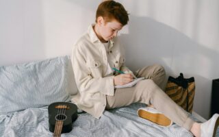 Preteen boy sitting on his bed writing on a piece pf paper.