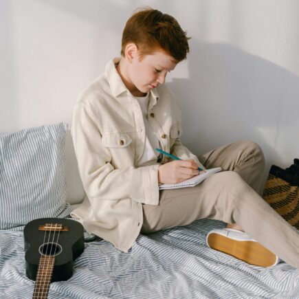 Preteen boy sitting on his bed writing on a piece pf paper.
