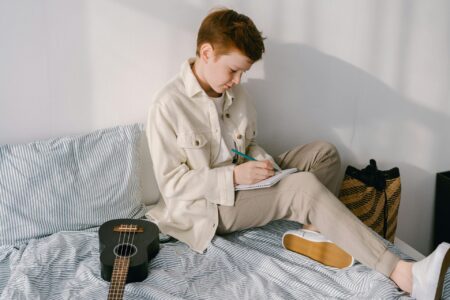 Preteen boy sitting on his bed writing on a piece pf paper.
