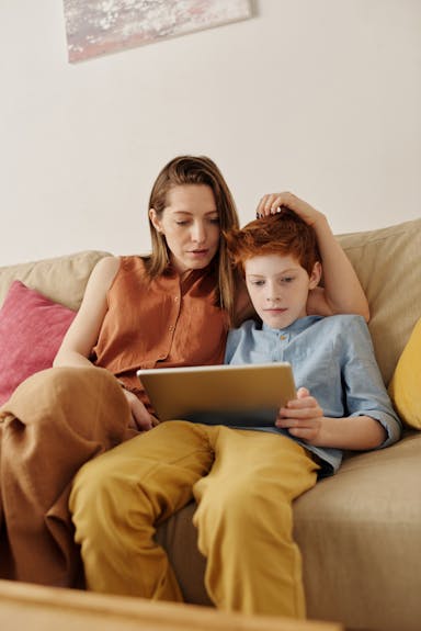 Mom sitting on a couch with her son. Mom is talking to him while he looks down at his tablet.