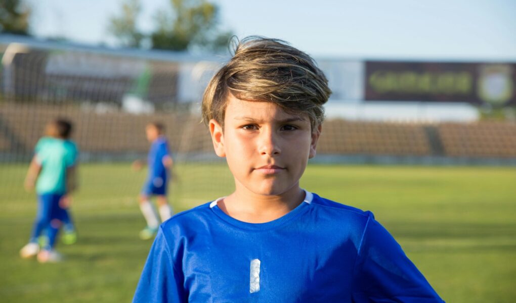 Boy standing in a soccer field looking at the audience, while 2 boys play in the back.