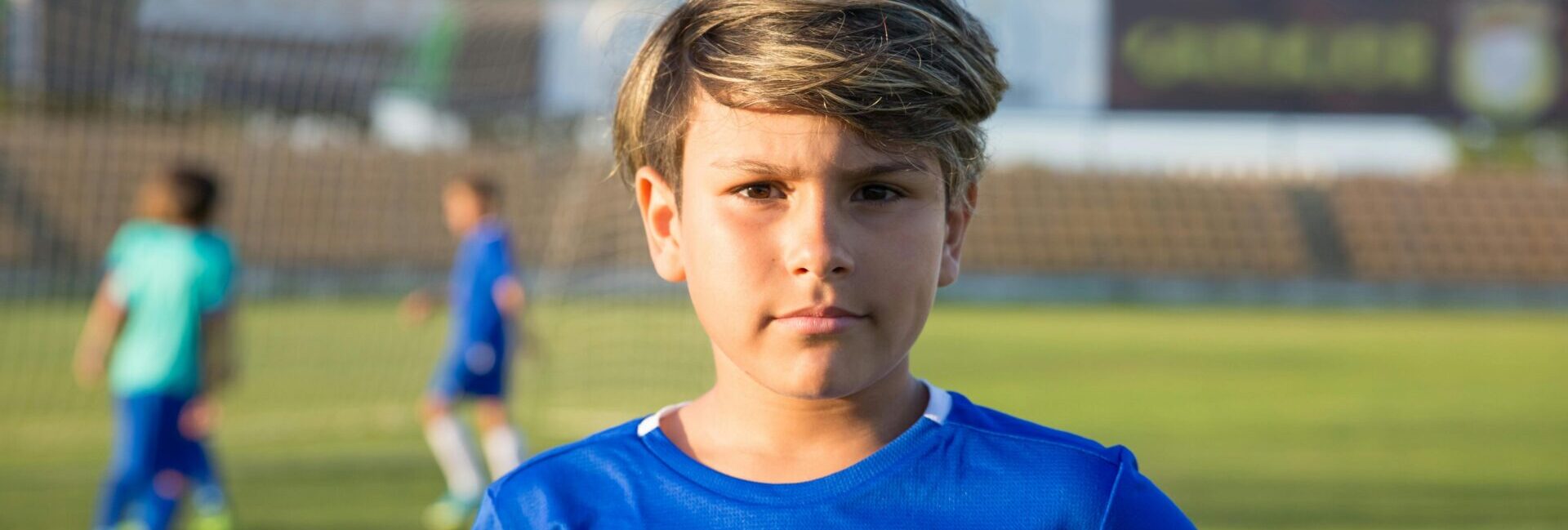 Boy standing in a soccer field looking at the audience, while 2 boys play in the back.