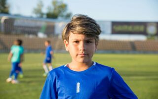 Boy standing in a soccer field looking at the audience, while 2 boys play in the back.