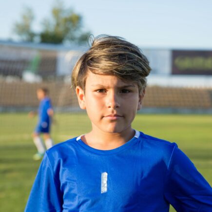 Boy standing in a soccer field looking at the audience, while 2 boys play in the back.