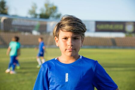 Boy standing in a soccer field looking at the audience, while 2 boys play in the back.