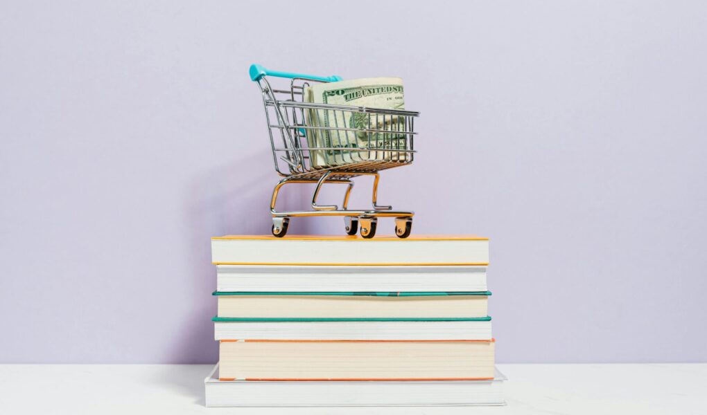 A small shopping cart with money in it stands on a book stack.