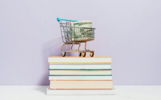 A small shopping cart with money in it stands on a book stack.