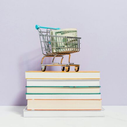 A small shopping cart with money in it stands on a book stack.
