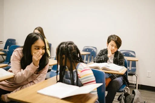 Two girls whisper to each other, one smiling. Behind them, a boy sits, looking uncomfortable. Peer pressure may move some kids to gossip about others.