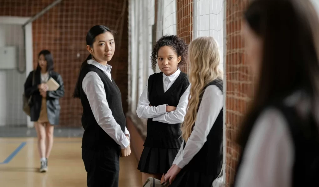 Three girls sit by a window talking with each other, while one of them is looking at another girl who is further away. This shows how peer pressure can exclude others from their peers.