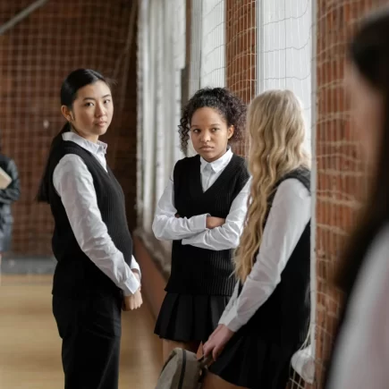 Three girls sit by a window talking with each other, while one of them is looking at another girl who is further away. This shows how peer pressure can exclude others from their peers.