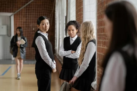 Three girls sit by a window talking with each other, while one of them is looking at another girl who is further away. This shows how peer pressure can exclude others from their peers.